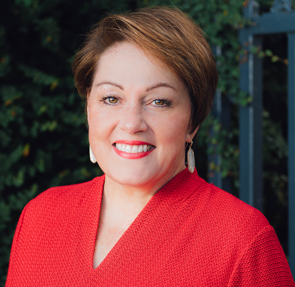 Smiling professional woman headshot in red textured jacket, short brown hair and drop earrings, outdoor background
