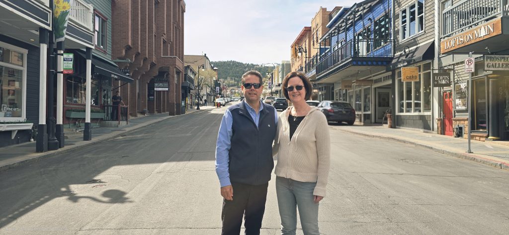 Couple smiling on a sunny small-town main street lined with shops, balconies, and parked cars