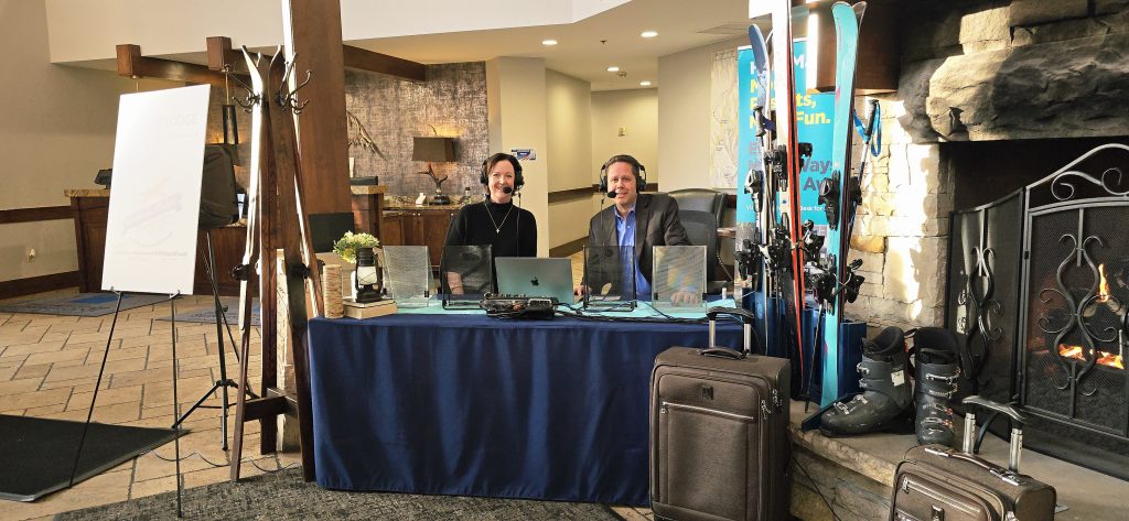 Two radio hosts broadcasting from a ski resort hotel lobby with laptops, skis, fireplace and luggage