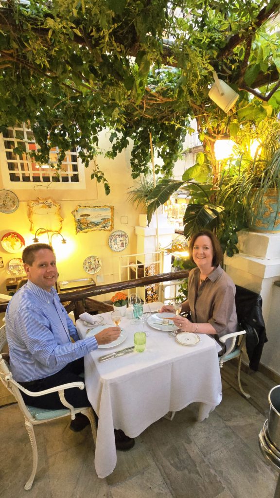 Couple enjoying wine at a white-tablecloth table under lush hanging greenery in a romantic terrace restaurant