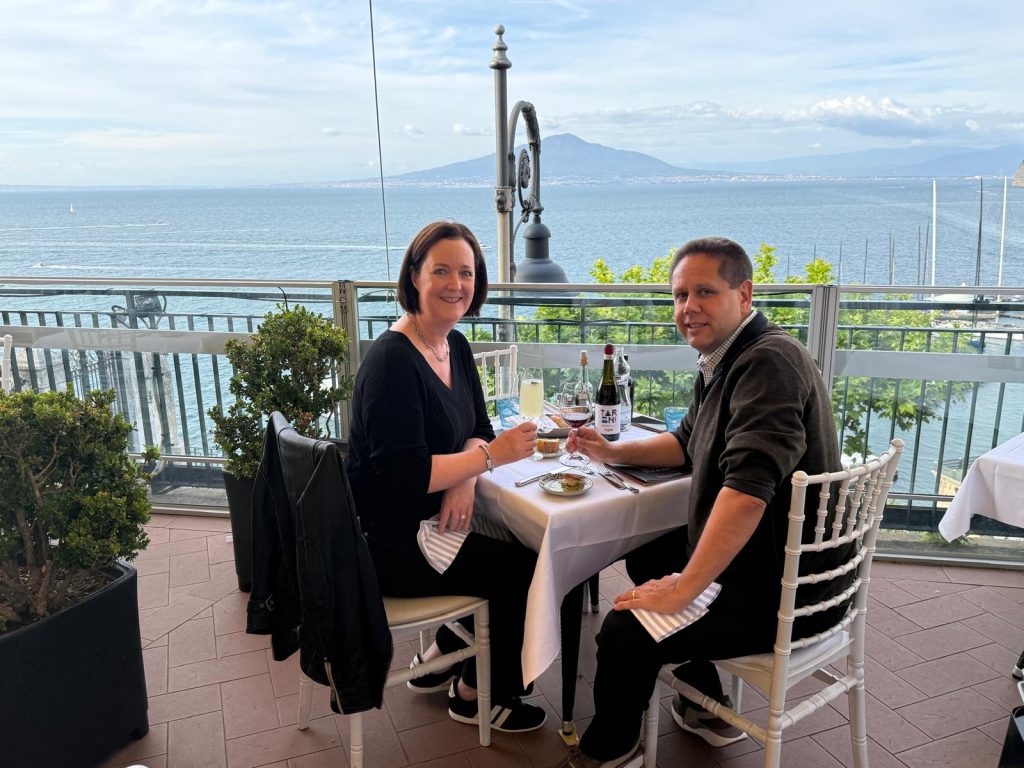 Couple enjoying a romantic dinner with wine on a seaside terrace overlooking the ocean and distant mountain backdrop.