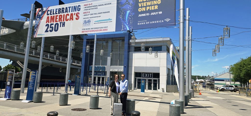 Intrepid Museum entrance with Celebrating America's 250th banner, couple with suitcase on pier under blue sky NYC