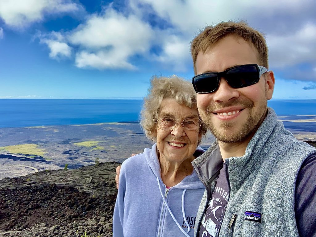 Smiling grandmother and adult grandson selfie on scenic rocky coastal overlook with blue ocean and cloudy sky
