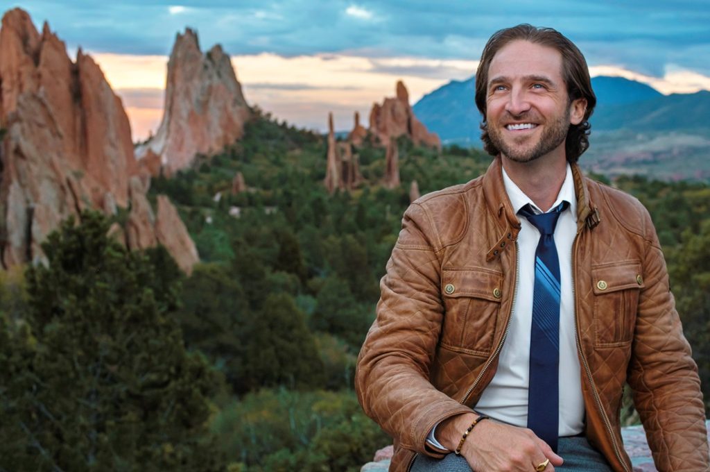 Outdoor portrait of smiling man in leather jacket and tie overlooking red rock formations and forested valley at sunset