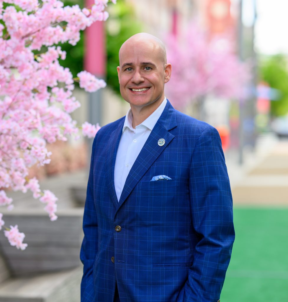 Professional headshot of smiling bald businessman in blue checked suit with pink cherry blossoms and blurred street