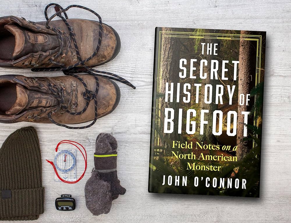 Bigfoot book The Secret History of Bigfoot with hiking gear — boots, compass, beanie on pale wood background