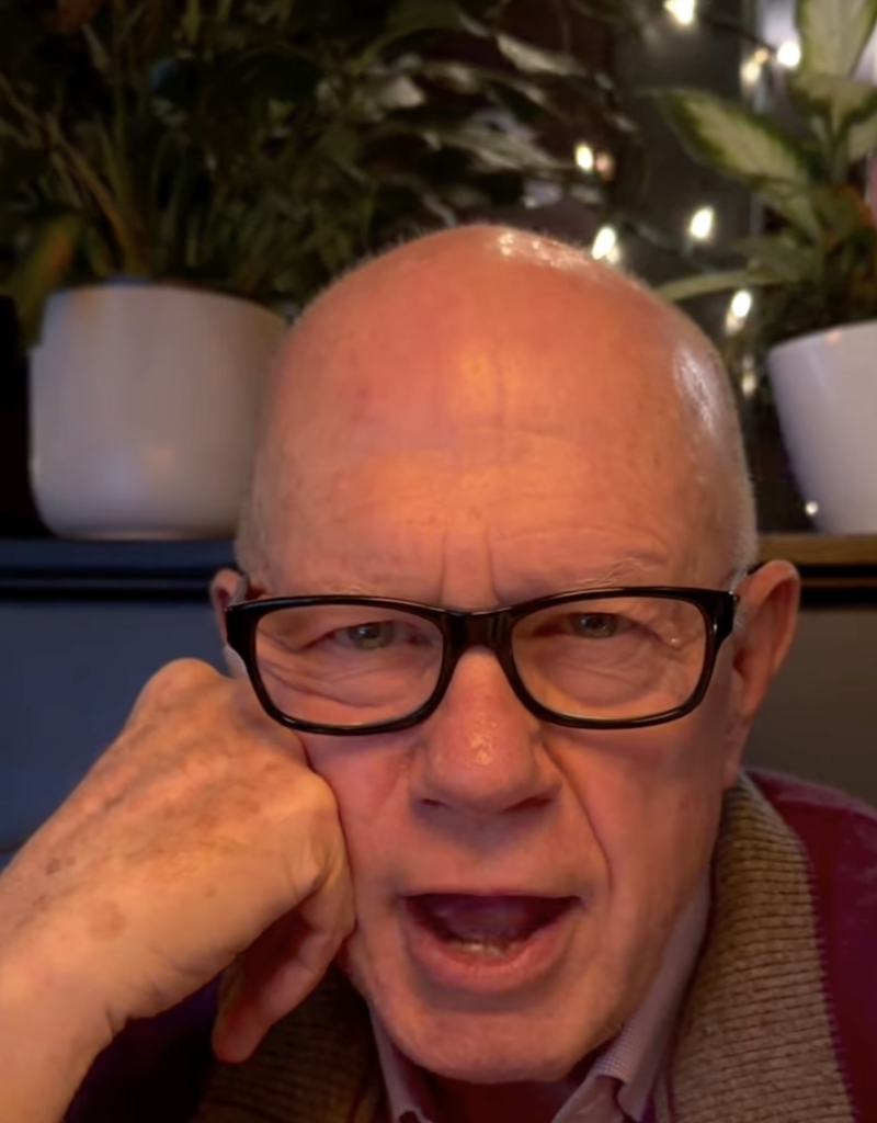 Close-up portrait of a bald older man in glasses speaking, hand on chin, plants in background, warm lighting