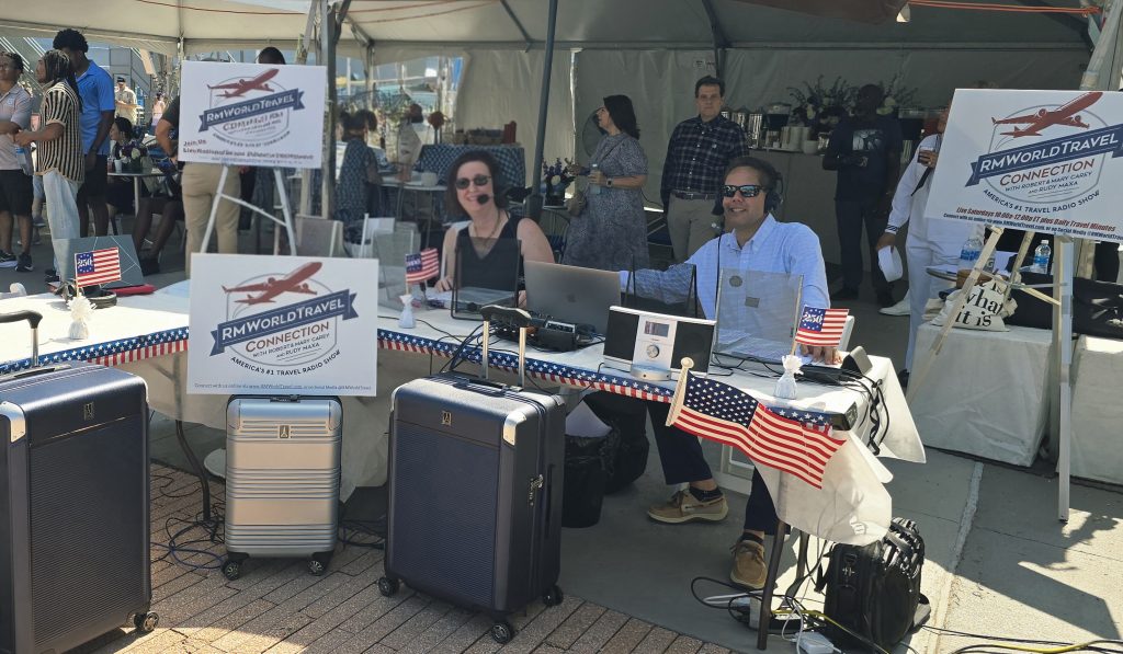 Travel radio show booth with hosts broadcasting, RMWorldTravel signage, luggage and American flags at outdoor festival