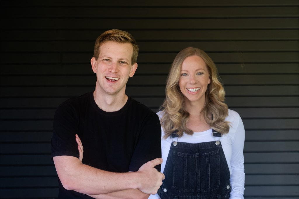 Smiling young couple portrait: man in black shirt and woman in white top with denim overalls, dark siding background