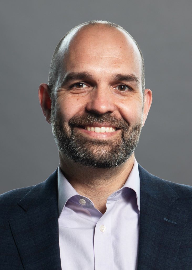 Corporate headshot: smiling bald bearded man in navy suit and light purple shirt on gray background